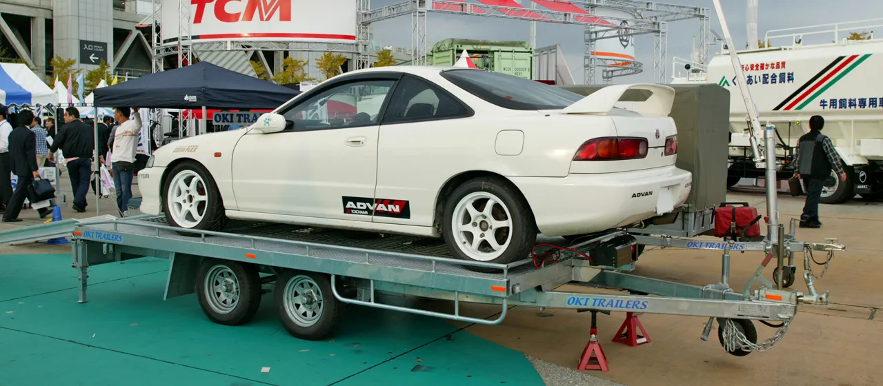 大型の2軸トレーラーに乗用車を積載した状態を斜め後方から見た写真。トレーラーはフラットな荷台とスチール製の枠を持ち、前端に連結用のカプラとジャッキが付いている