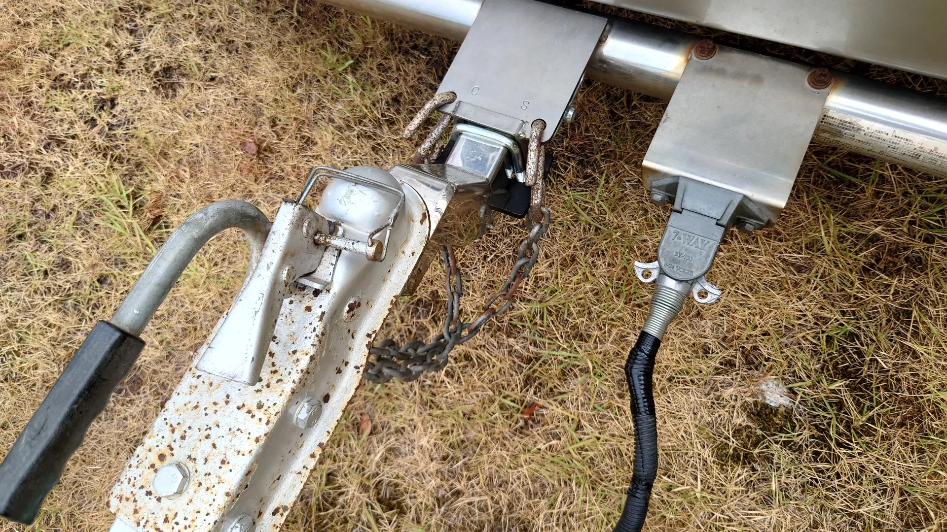 トレーラー前端のカプラを牽引車のヒッチボールに掛けた状態の接写。カプラの脇から2本の安全チェーンがヒッチメンバーに掛けられ、右側には灯火用の電気接続コネクタが差し込まれている