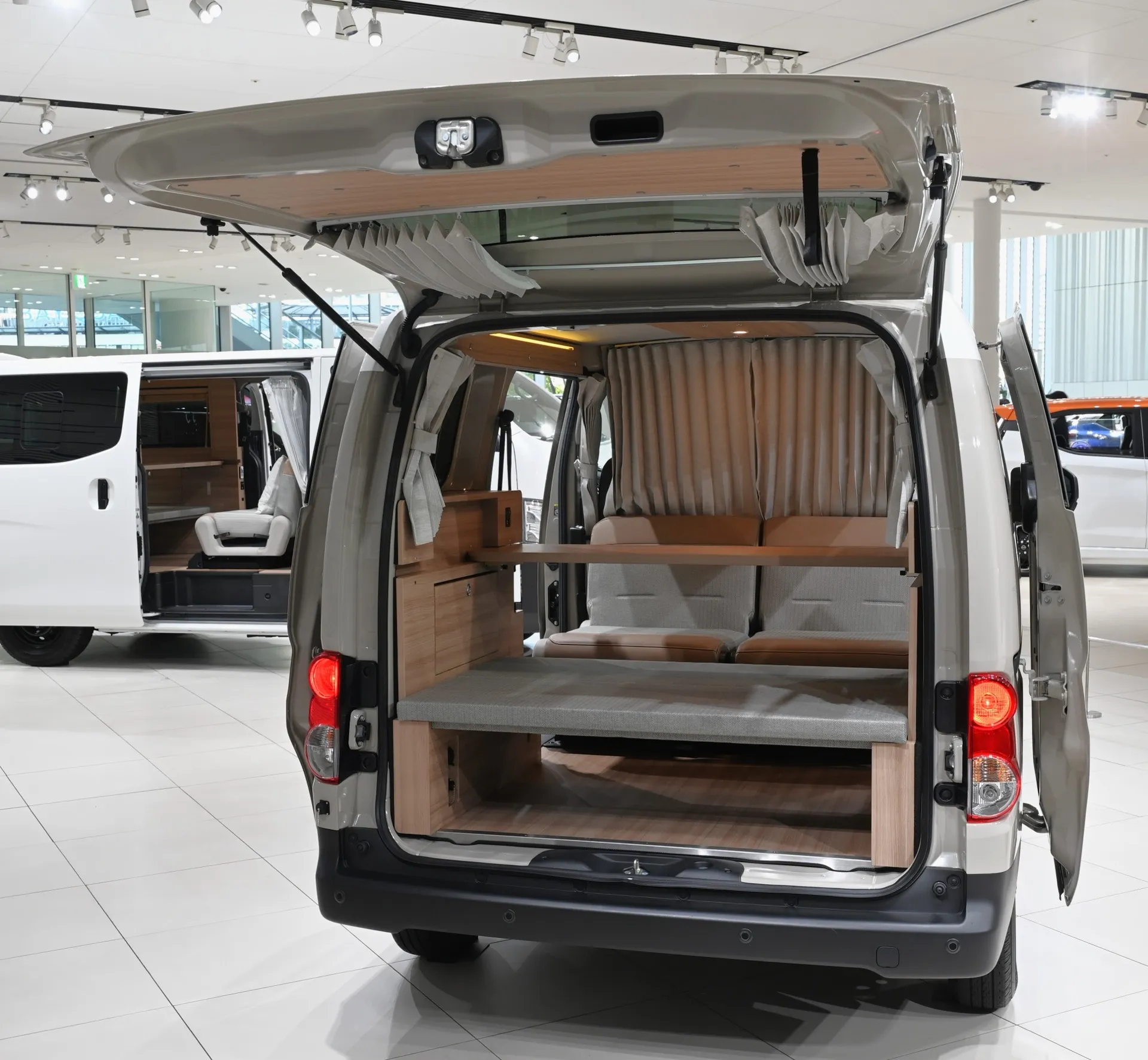 NV200バネットの車中泊仕様の荷室。木目調のフロアとサイドトリム、平置きベッド、車内カーテンが並ぶ（2024年12月撮影）