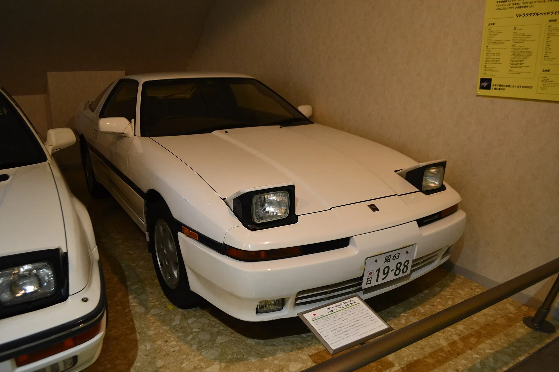 白いトヨタ スープラ1代目を博物館の展示室で右斜め前から見た外観。リトラクタブル式ヘッドライトが上がった状態で、バンパーの右端に「TWIN TURBO」の文字が入る
