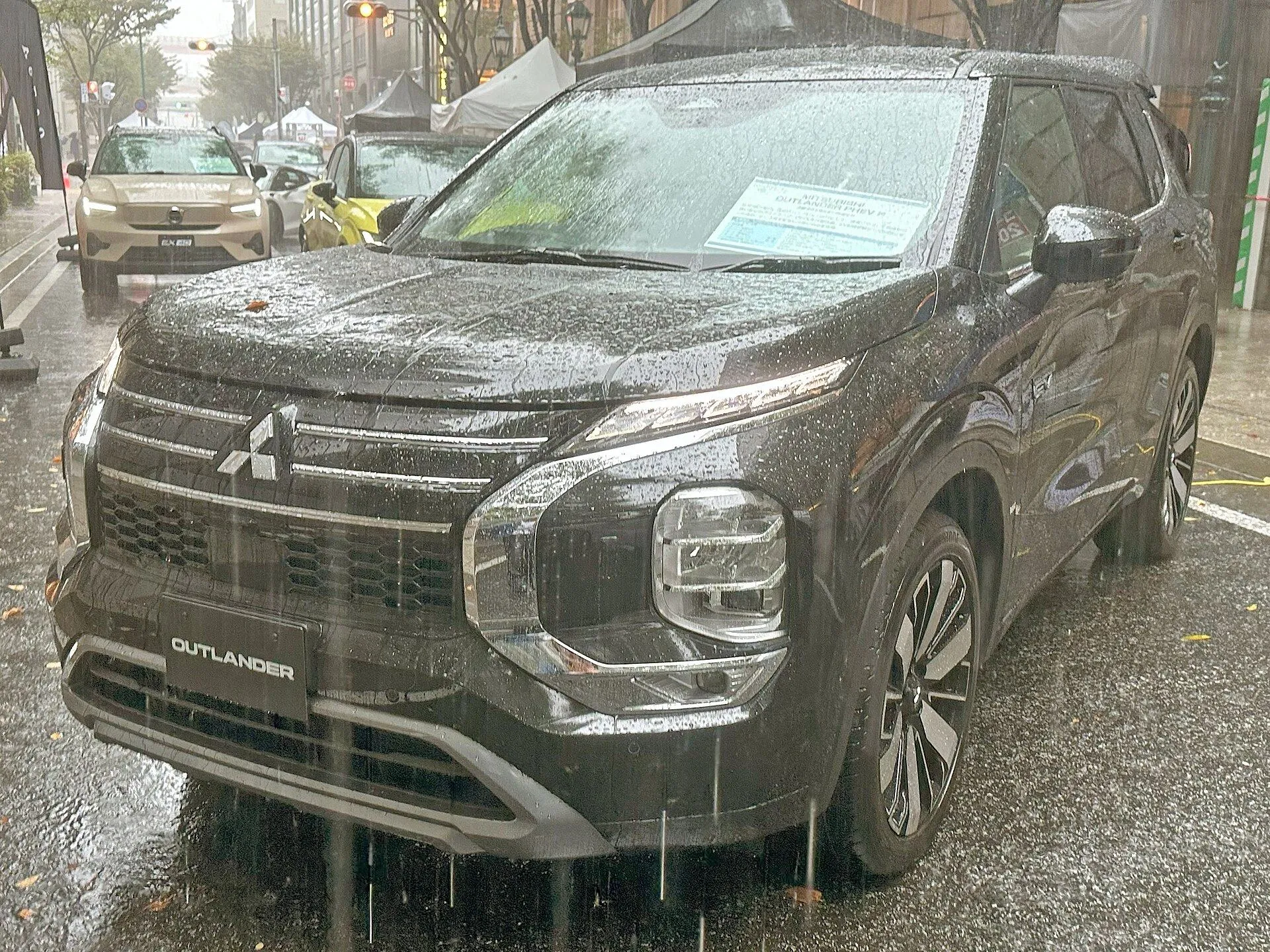 雨の路上に停まる濃色の三菱 アウトランダーPHEV P（5LA-GN0W）を斜め前から見た外観