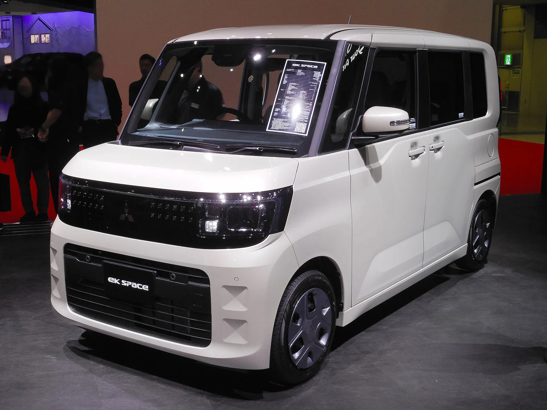 白い三菱 eKスペース G（2025年式）の展示車を左前方から見た外観。横長のヘッドライトとグリル、ホイールカバー付きのホイール、助手席側の2枚のドアが見える