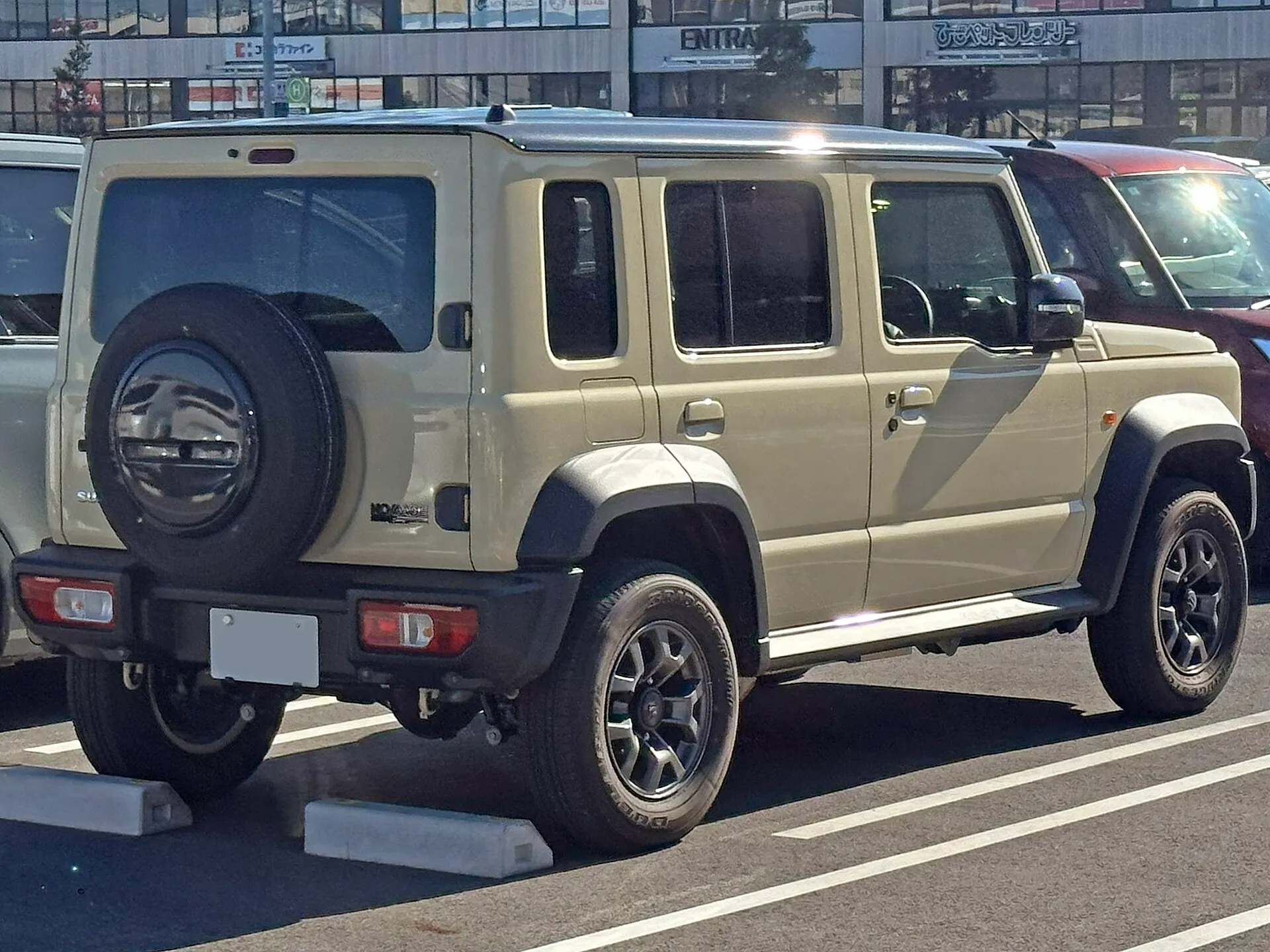 駐車場に停まったスズキ ジムニー ノマド FC（3BA-JC74W）を斜め後ろから見た外観。明るいベージュ系のボディに黒い屋根で、バックドアにスペアタイヤを背負い、側面に前後2枚のドアが並ぶ