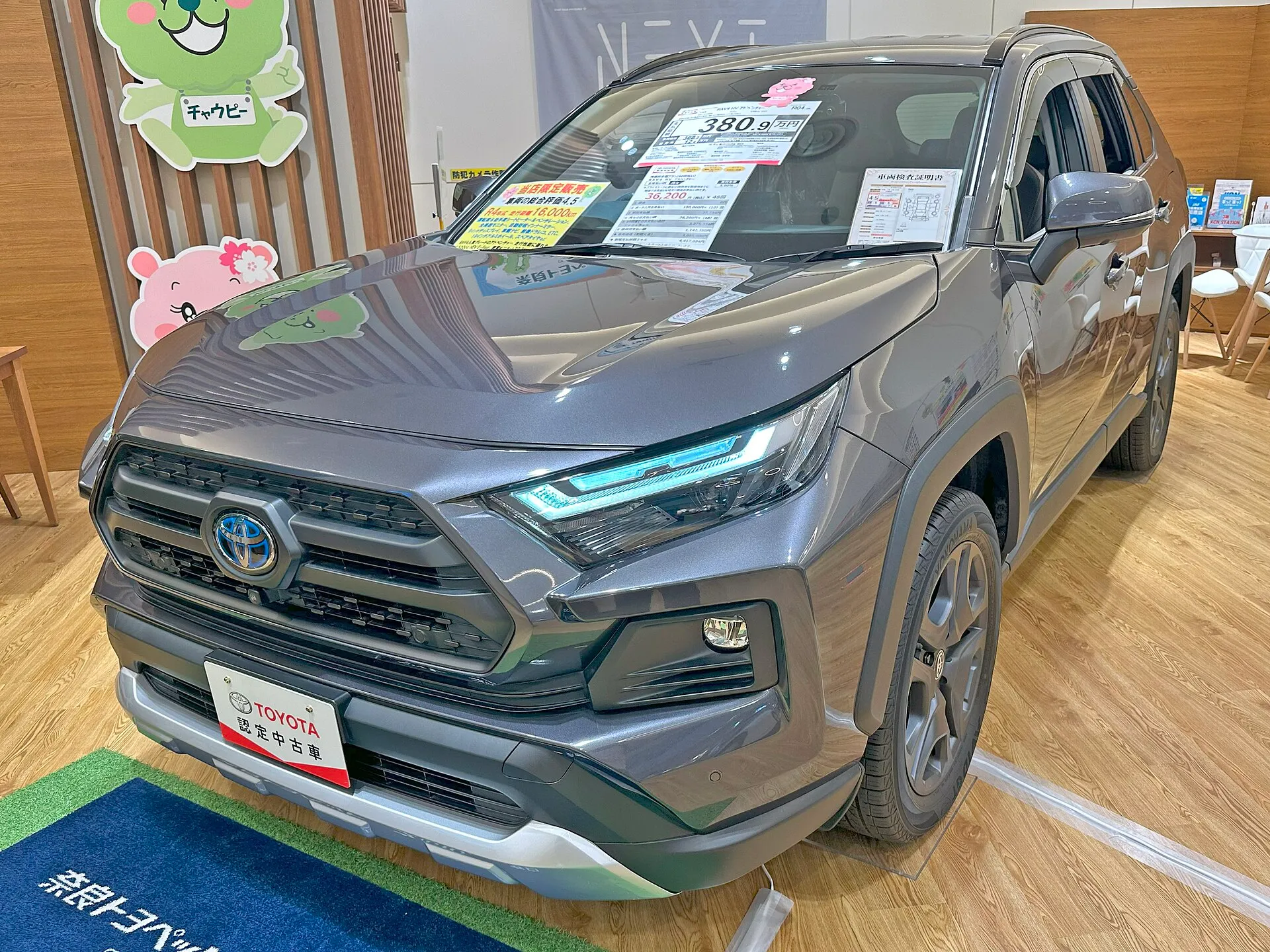 濃いグレーのトヨタ RAV4 HYBRID Adventureを左前方から見た外観。屋内の展示スペースに置かれ、グレーのアルミホイールを履いた、型式6AA-AXAH54-ANXVBの個体