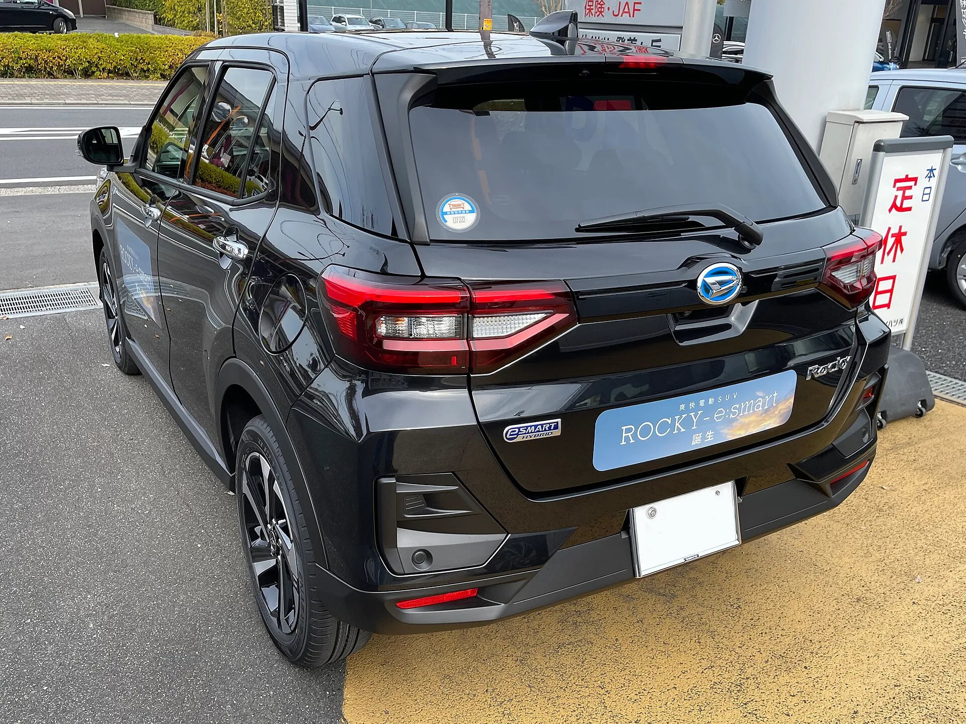 黒いダイハツ ロッキー Premium G HEVを左斜め後ろから見た外観。販売店の駐車場に置かれ、赤いテールランプと、リヤに付けた「ROCKY-e:smart」の表示、メッキのドアハンドルが見える、型式5AA-A202S-GBSHの個体