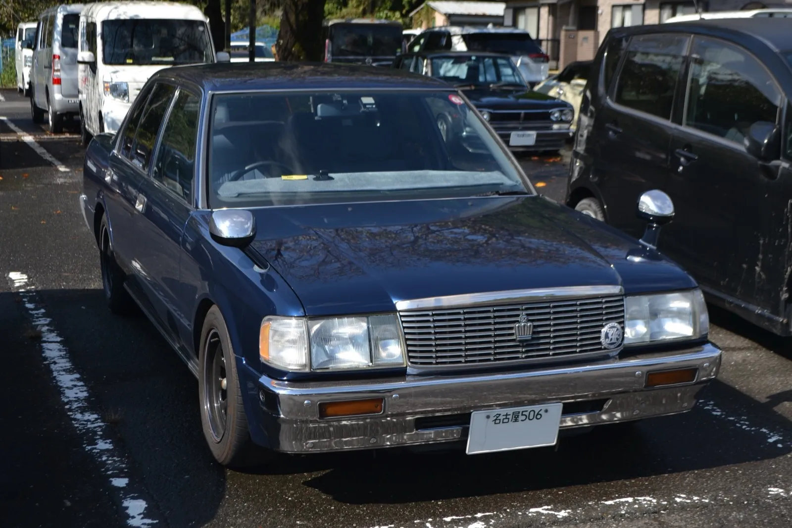濃紺のトヨタ クラウン セダン デラックス（型式E-GS130）を右斜め前から見た外観。角形4灯のヘッドライトと縦桟のグリルが並び、車体側面に細いモールが1本付いている