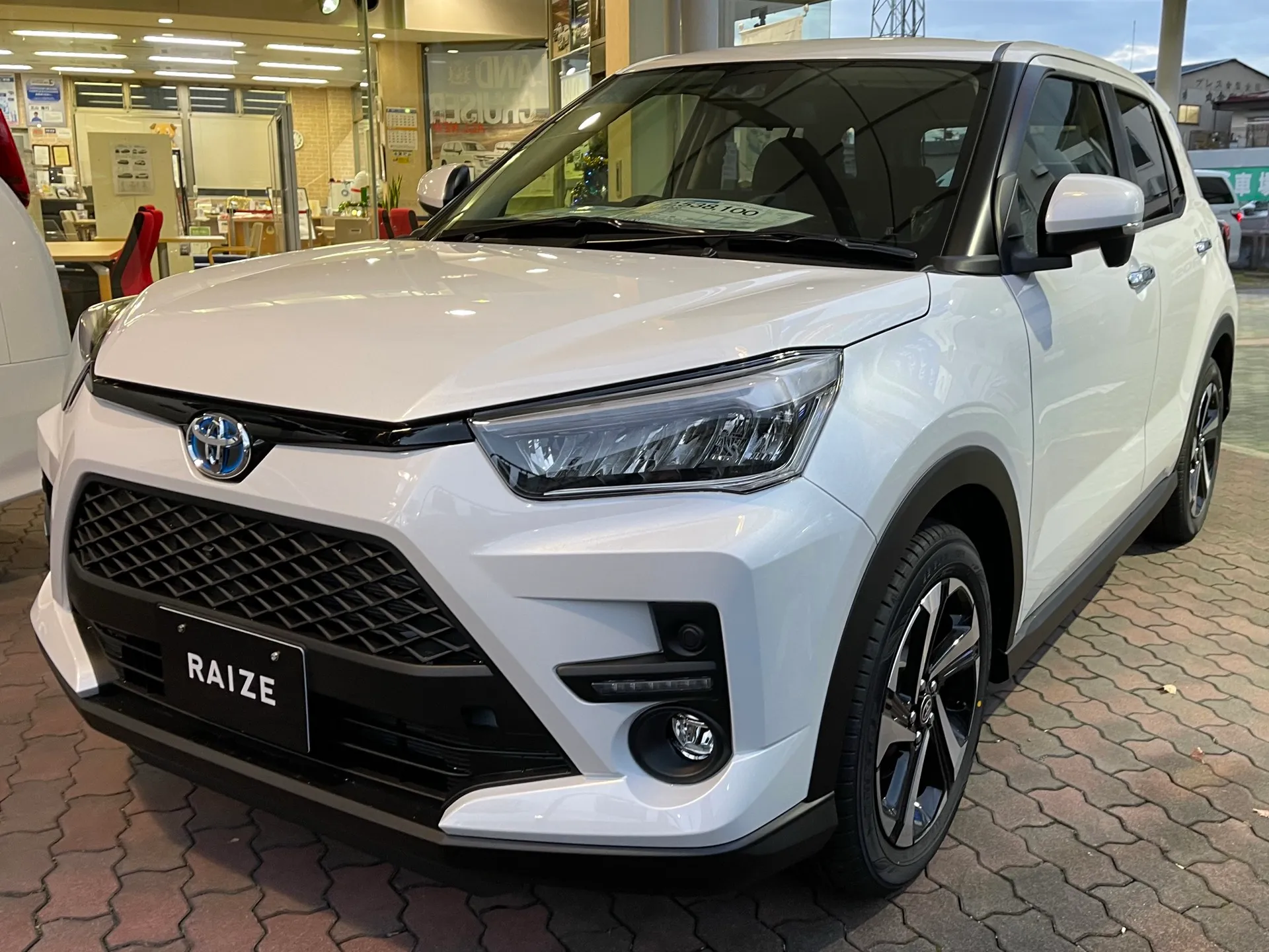 白いトヨタ ライズ HYBRID Z（型式5AA-A202A-GBSH）を左斜め前から見た外観。販売店のショールームの前に置かれ、フロントバンパーの丸いフォグランプと切削加工のアルミホイールが見える