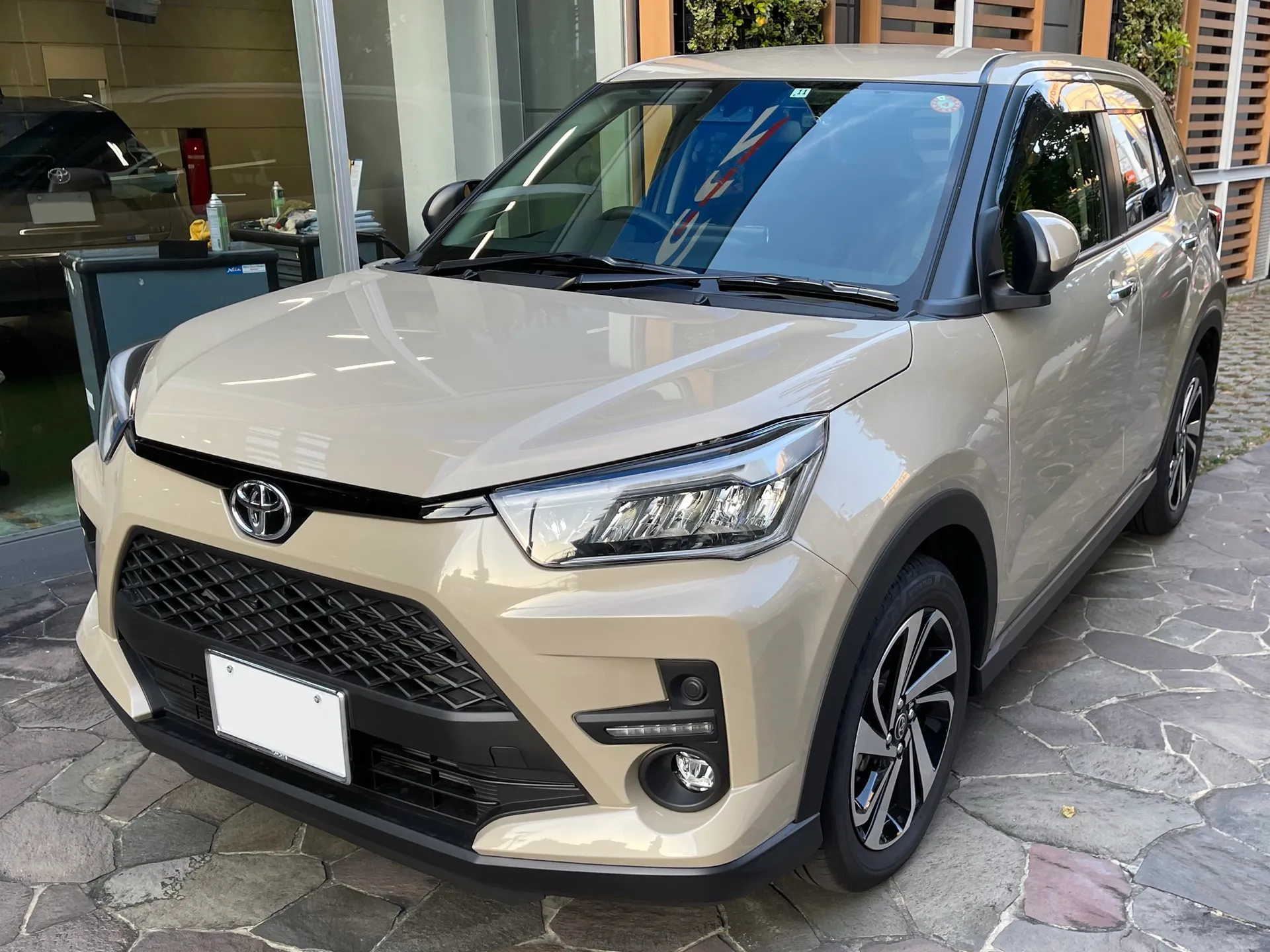 ベージュのトヨタ ライズ Z 2WD（型式5BA-A201A-GBSF）を左斜め前から見た外観。販売店の石畳に置かれ、細いLEDヘッドランプ、バンパー下のフォグランプ、切削光輝とブラック塗装のアルミホイールが見える