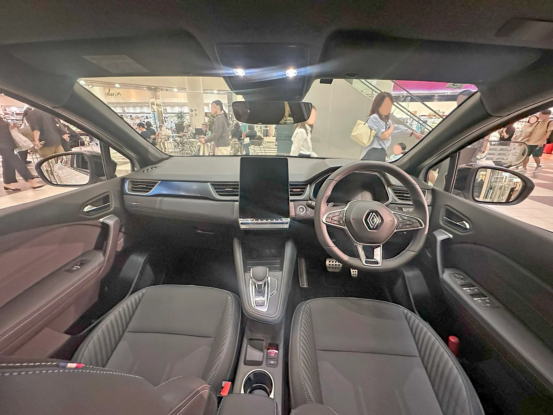 ルノー キャプチャー エスプリ アルピーヌ マイルドハイブリッド（型式3AA-HJBH5HH）の運転席まわりを後席側から見た写真。右ハンドルで、中央に縦長の画面、足元にアルミ製のペダル、天井から縁のないルームミラーが下がっている。前席は濃いグレーの表皮