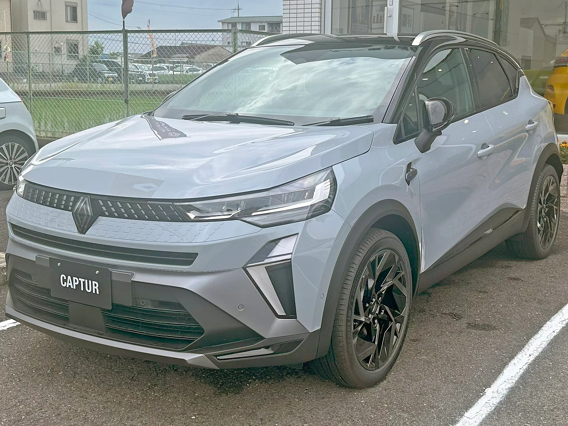 駐車場に置かれた、青みのあるグレーの車体に黒いルーフのルノー キャプチャー エスプリ アルピーヌ フルハイブリッド E-TECH（型式5AA-HJBH4MH）を斜め前から見た外観。ナンバーの位置にCAPTURと書かれたプレートが付き、ルーフの端に銀色のレールが見える