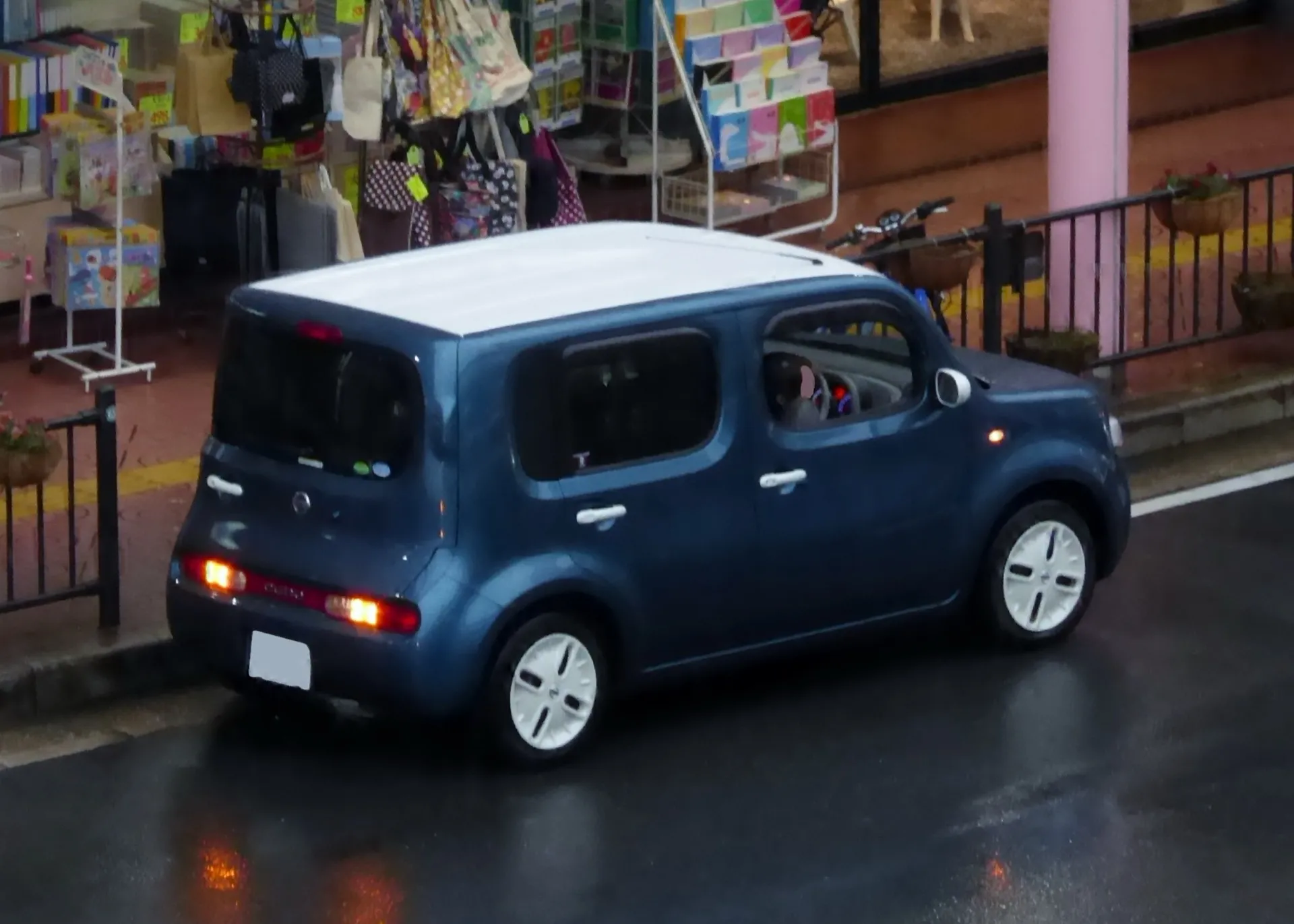 日産 キューブ（Z12）を左後方から見た外観。青いボディに白いルーフと白いフルホイールカバーの2トーンカラーで、リヤウインドウが左側へ回り込んでいる