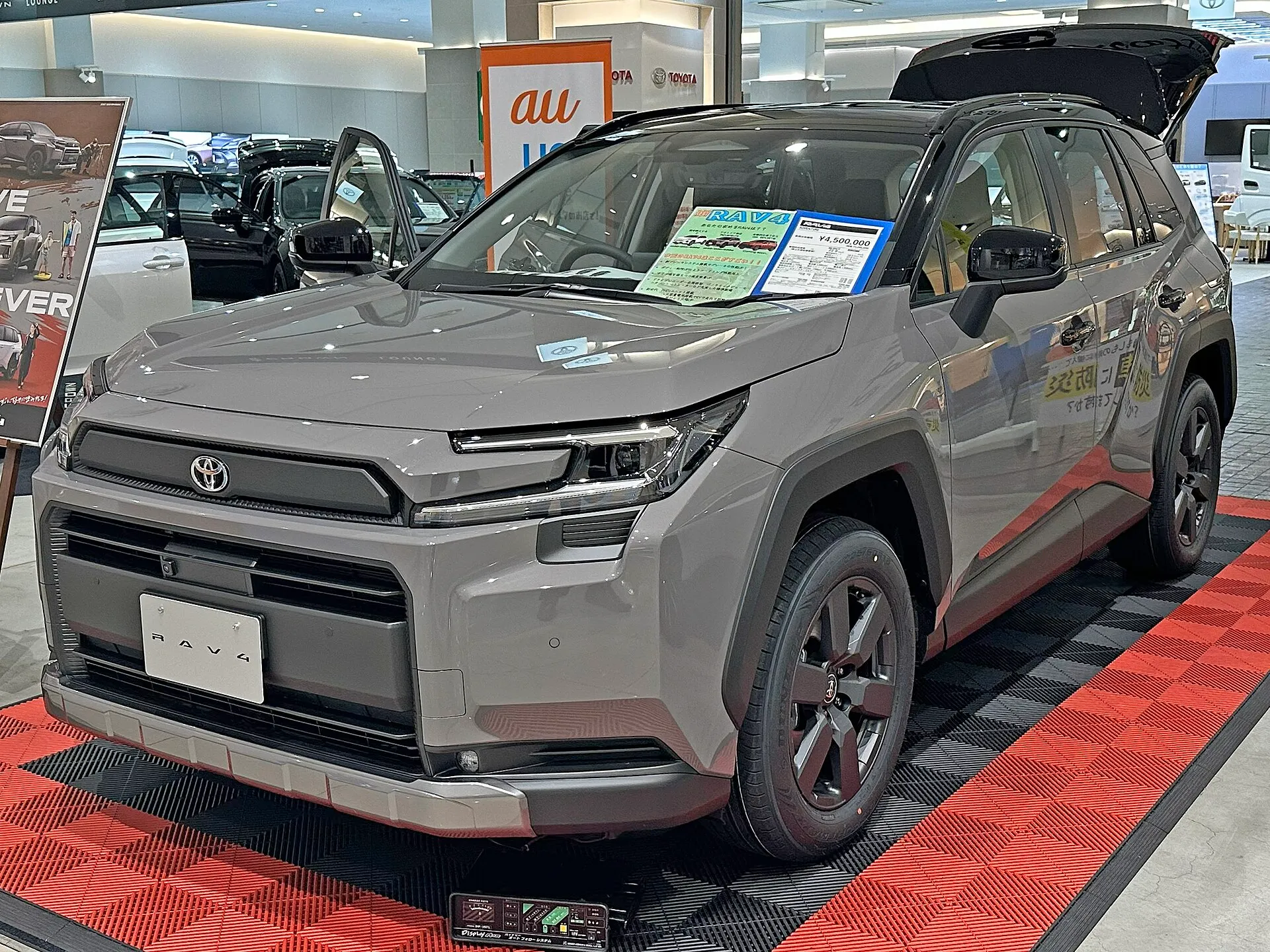 トヨタ RAV4 Adventure（ハイブリッド車・6AA-AXAN64-ANXVB）を左斜め前から見た外観。グレーのボディに黒い樹脂のフェンダーアーチを持ち、バックドアを開けた状態で屋内の展示スペースに置かれている