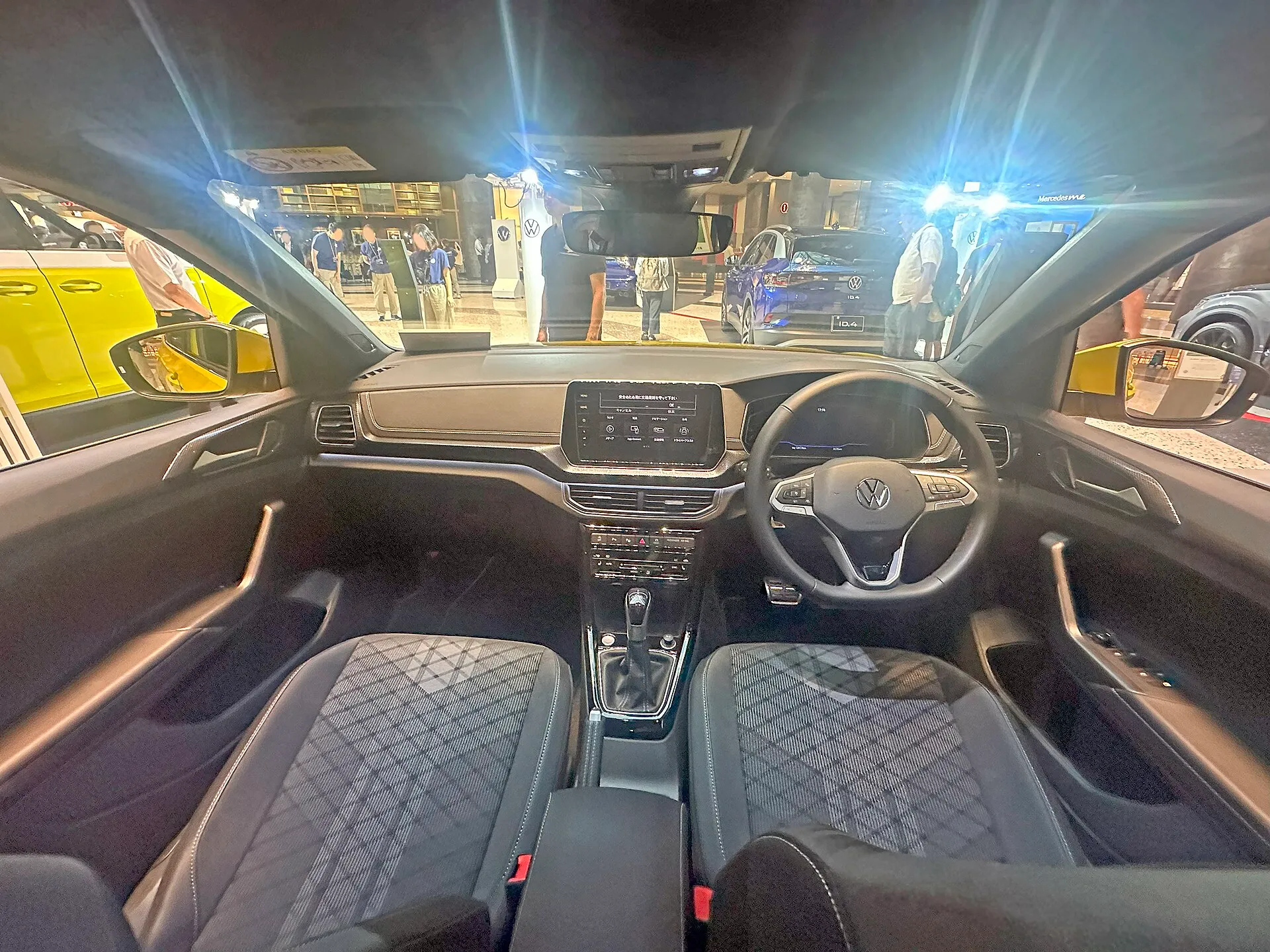フォルクスワーゲン T-Cross TSI R-Line（型式3BA-C1DKR・右ハンドルの日本仕様）の前席と運転席まわり。ダッシュボード中央に横長の画面があり、その下に空調の操作パネル、手前にシフトレバーが並ぶ。屋内の展示会場で撮影された個体