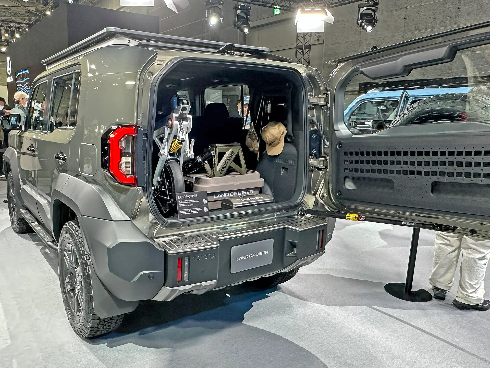 横開きのバックドアを開けた、くすんだ緑色のトヨタ ランドクルーザー“FJ”（TRJ240）を左斜め後ろから見た外観。バックドアは後ろから見て右側のヒンジで横に開き、荷室には折りたたみ式の小型モビリティと収納ボックスが積まれている。屋根にルーフラック、バックドアの内側に穴の開いたパネルが付いた展示車