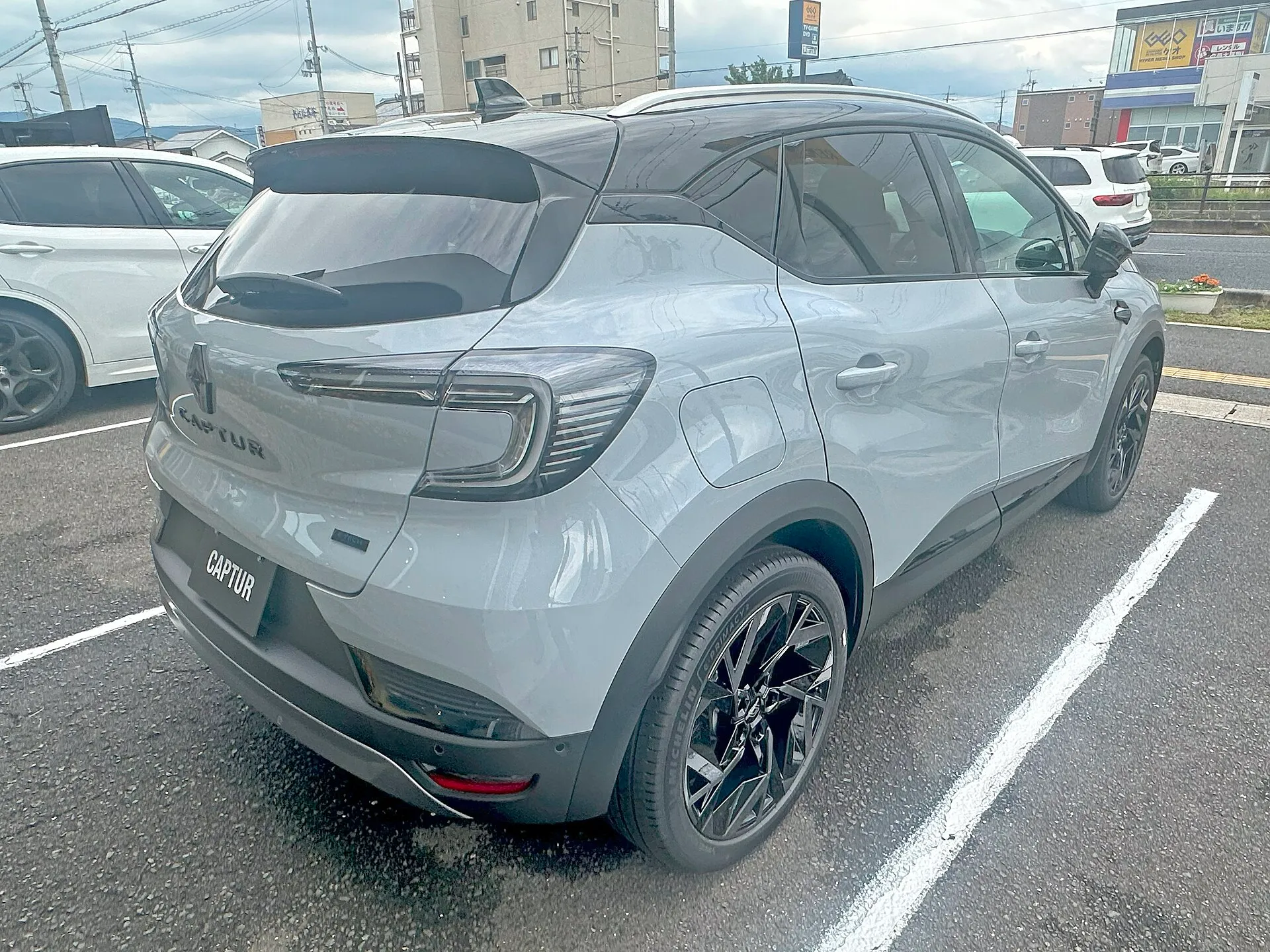 駐車場に置かれた、青みのあるグレーの車体に黒いルーフのルノー キャプチャー エスプリ アルピーヌ フルハイブリッド E-TECH（型式5AA-HJBH4MH）を斜め後ろから見た外観。バックドアにCAPTURの文字があり、ナンバーの位置にもCAPTURと書かれたプレートが付く