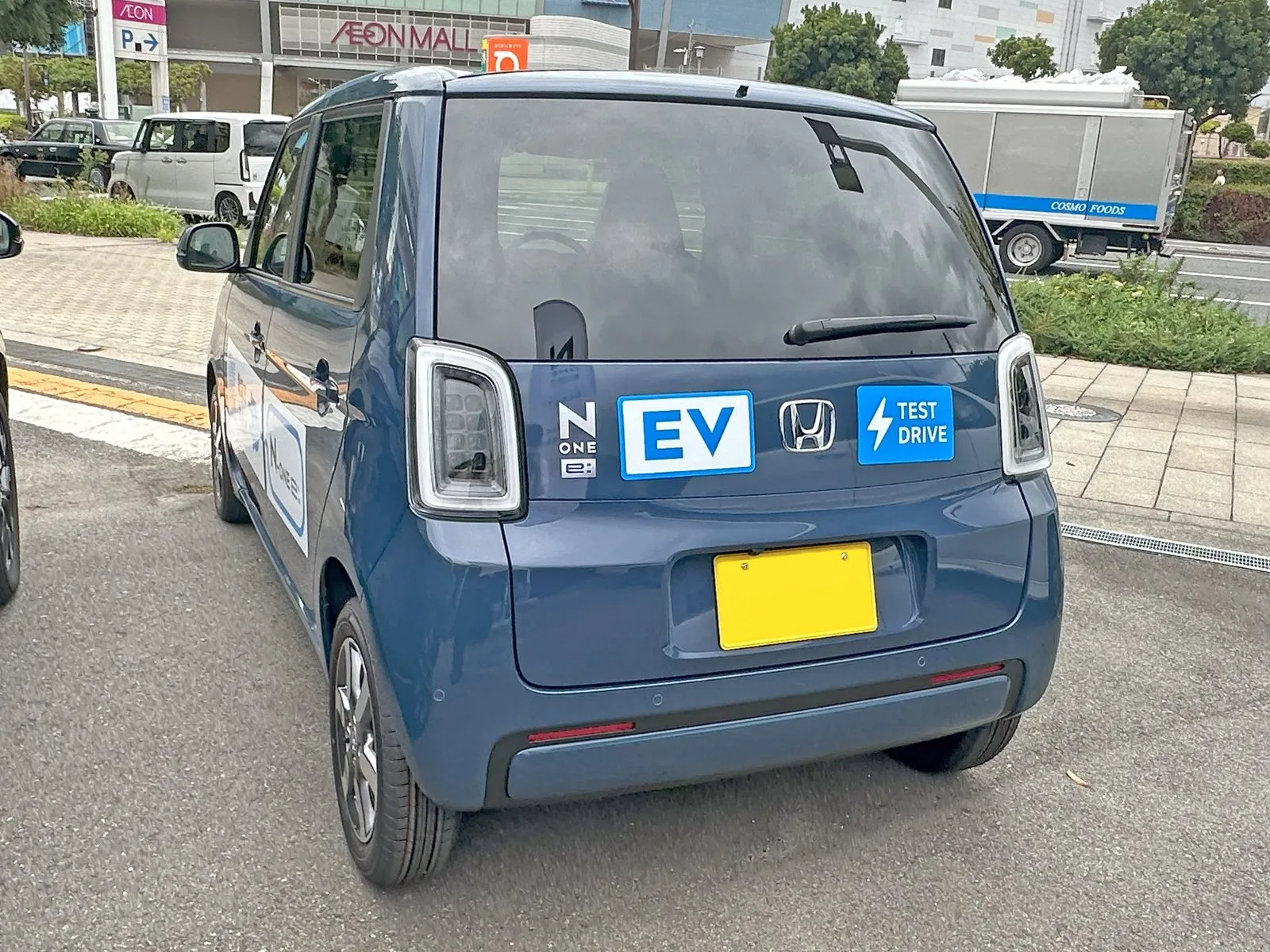 ホンダ N-ONE e: の e: L（ZAA-JG5）を斜め後ろから見た外観。青い車体で、バックドアに N-ONE e: のエンブレムと EV・TEST DRIVE と書かれたステッカーがあり、黄色いナンバープレート（番号は写っていない）が付いている