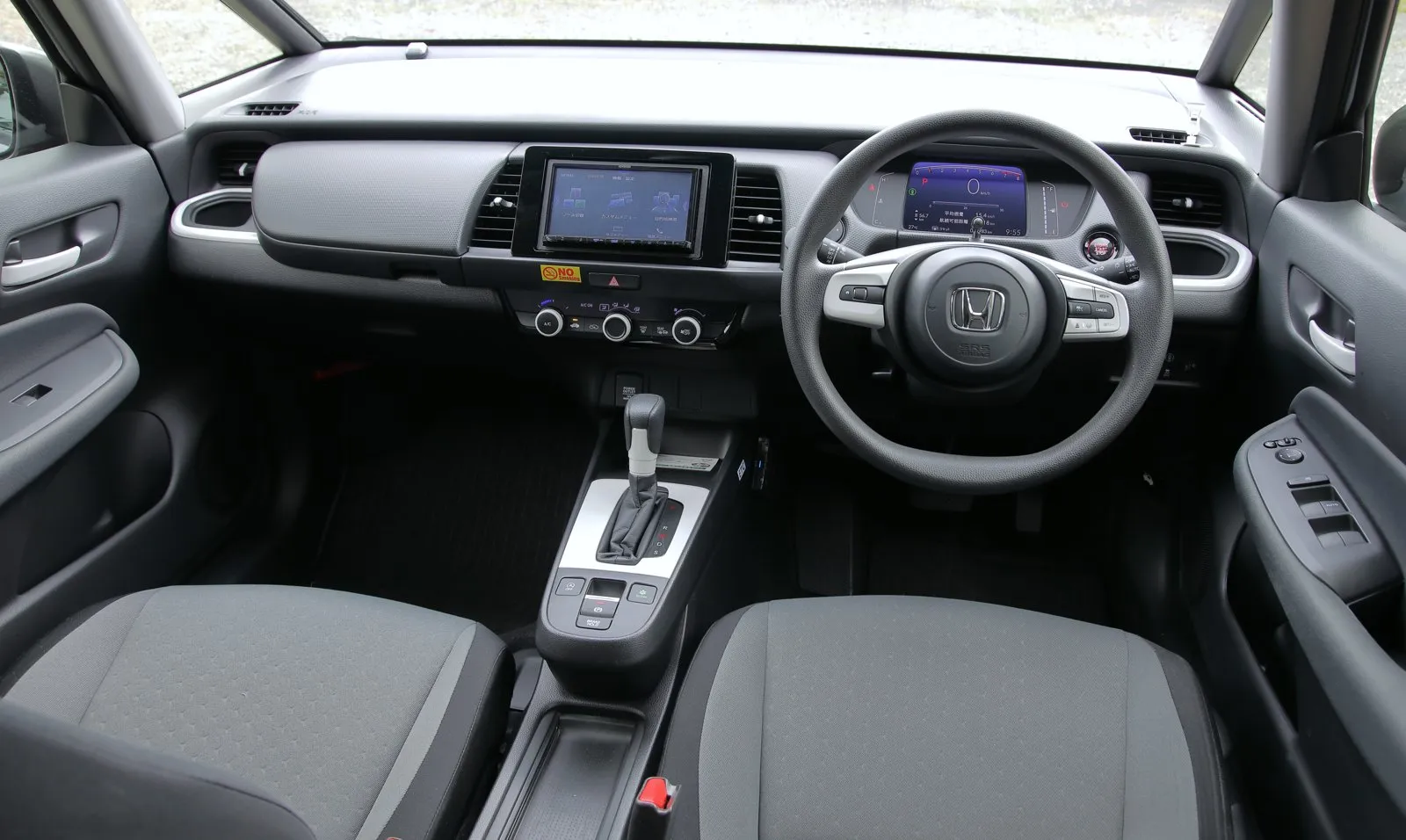 ホンダ フィット BASIC 4WD（6BA-GR2）の運転席まわりを、後席のあたりから見た写真。2026年7月のマイナーモデルチェンジより前の個体で、BASIC はこの改良で名前が X に変わった。前席の間はひじ掛けのない低いトレーで、エアコンの操作部は3つのダイヤル。ステアリングホイールは2本スポークで、シートは灰色の布張り。中央にナビの画面が付いている